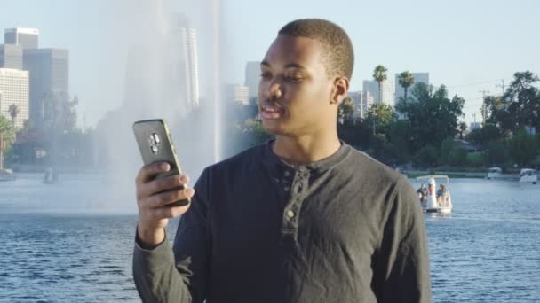 Joven hombre afroamericano usando su teléfono, mirando a su alrededor frente al lago — Vídeo de stock