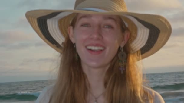 Jeune femme avec un chapeau souriant à la caméra pendant le coucher du soleil près de l'océan — Video