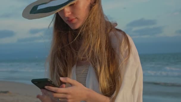 Mulher bonita usando seu celular pelo oceano durante o pôr do sol — Vídeo de Stock