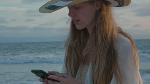 Молода жінка на пляжі смс біля океану під час заходу сонця — стокове відео
