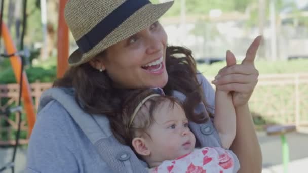 Žena se usmívá, zatímco drží dítě v nosítkách na hřišti — Stock video