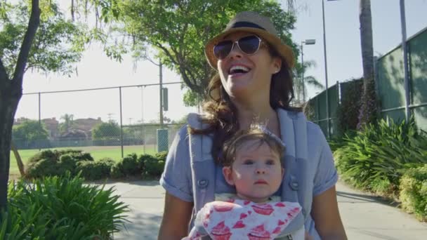 Женщина с ребенком в носильщике улыбается, когда идет в парк — стоковое видео