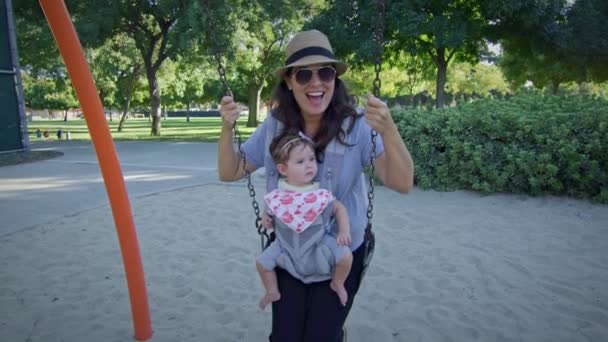 遊び場でキャリアの赤ちゃんとスイングに揺れる女性 — ストック動画