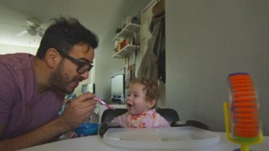 Baba 'yı kaşıkla beslerken bebek kızıyla oynarken çok güzel bir kare.