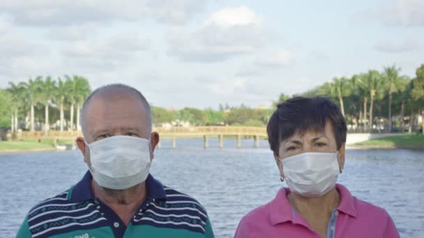 Älteres Paar blickt in Kamera, während es sich vor dem See die Gesichtsmasken auszieht — Stockvideo