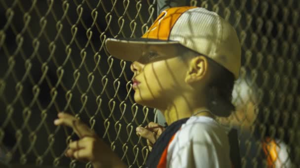 Young boy standing by fence in dugout looking towards baseball game — Stock Video