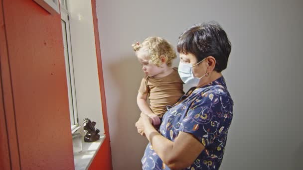 Abuela usando mascarilla, sosteniendo al bebé y mirando por la ventana — Vídeo de stock