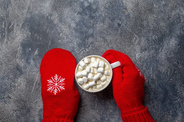 Natal e Ano Novo composição de férias acolhedora com cachecol, mãos de mulher em mitenes, canecas com bebida quente e marshmallow no fundo de concreto cinza. Deitado plano, vista superior. — Fotografia de Stock