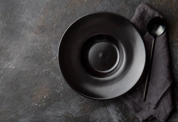 Fondo alimentare con piatto vuoto nero, tovagliolo grigio e cucchiaio nero su sfondo scuro — Foto Stock