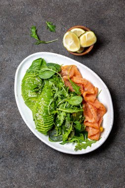 Az karbonhidratlı salata. Ispanak, roka salatası, avokado ve somon. Siyah beton arka plan, beyaz plaka, üst manzara