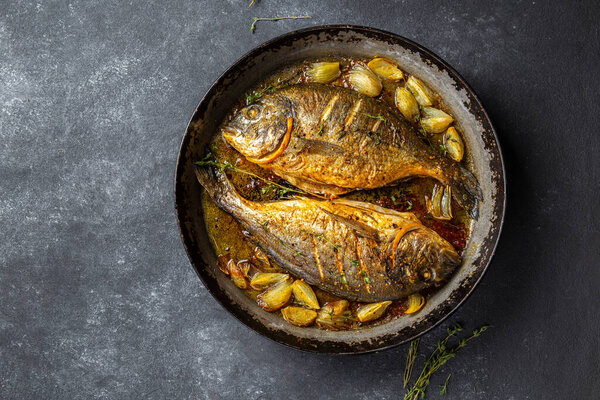 Baked sea bream or dorada with onion and herbs in pan on dark background.
