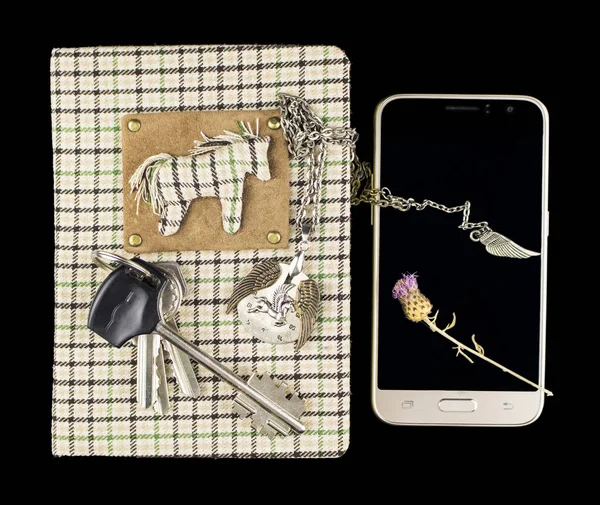 A handmade notebook with a checkered surface on which lies a bunch of keys and a pendant on a chain in the form of a dial with a pegasus and a number of smartphones on which lies a dry thistle flower, on a black background