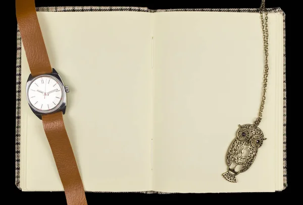 A vintage handmade notepad unfolded on empty yellowed pages, with an antique wristwatch and a pendant on a chain in the form of an owl, on a black background