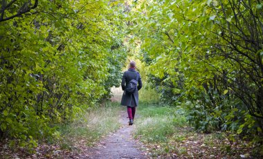 A path through thickets of bushes and trees, along which comes the girl in a long green coat, red pants and with a backpack on her shoulder clipart
