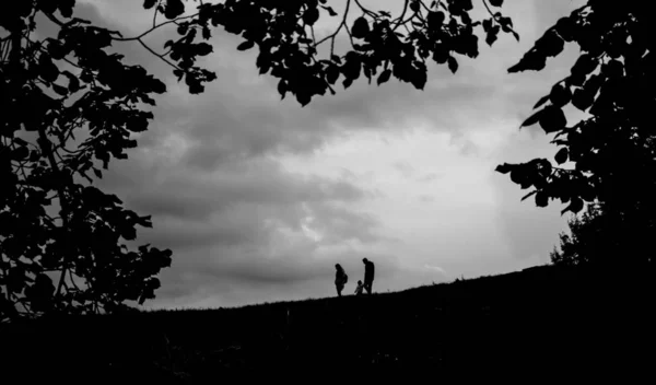 Black silhouette of a slope from which a family of three people descends on the background of the sky and framed by leaves