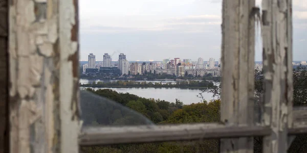 在一座高楼高耸的城市、一条长满绿岛的河流和高高的树梢上，从一个破了的旧窗户眺望 — 图库照片