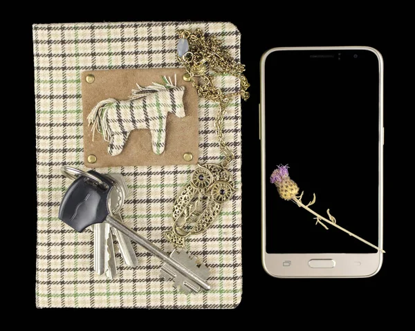 A handmade notebook with a checkered surface on which there is a bunch of keys and a pendant on a chain in the form of an owl and next to the smartphone on which lies a dry thistle flower, on a black background