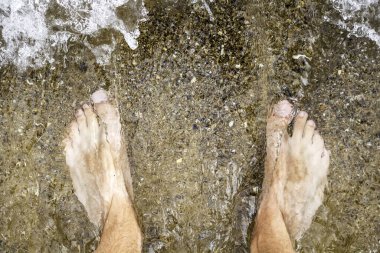 Erkek bacakları suyun altında, çakıl taşları şeffaf bir dalga aracılığıyla görülebilir.