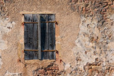 Kısmen tahrip olmuş alçıyla kaplı eski bir tuğla duvar parçası. Kararmış ahşap panjurlarla kaplı bir pencere var. Doku. Arkaplan.