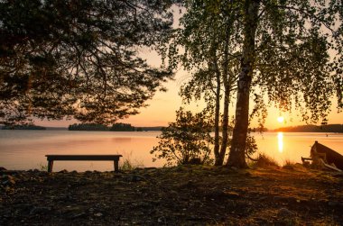 Büyük bir göl üzerinde gün batımı manzarası. Kıyıdaki ağaçların, kayıkların ve bankların siluetleri görülebiliyor. Arka planda uzak bir kıyı, batan güneş ve suyun üzerinde parlak bir yol var. Peyzaj