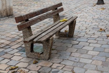 Taş döşeli bir caddede eski bir bank duruyor. Beton bir taban ve tahta bir koltuk var. Asfaltta birkaç sonbahar yaprakları uzanıyor. Geçmişi. Peyzaj.