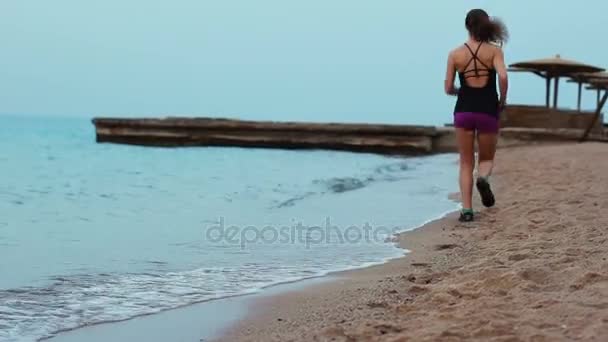 Sportowe dziewczyna biegnie wzdłuż piaszczystej plaży, na którym toczą się fale. Brunetka robi rano bieganie na wybrzeżu. — Wideo stockowe