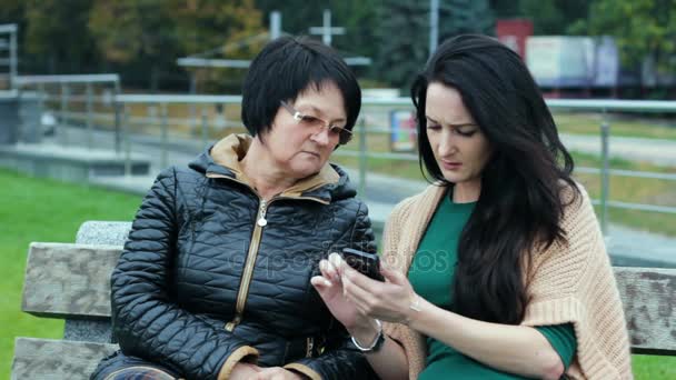 The younger generation explains to the elder how to use modern gadgets. Two brunettes of different ages are sitting on a bench. — Stock Video