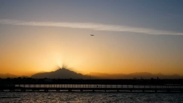 O avião voa sobre o pôr do sol sobre as montanhas. Raios de luz passam pela névoa da noite. Mar calmo e cais em primeiro plano . — Vídeo de Stock