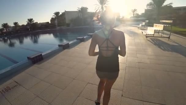 Ein schlankes athletisches Mädchen läuft morgens in der Nähe des Pools gegenüber der Sonne. Helle Strahlen durchdringen die Haare. Langsames Motiv. — Stockvideo