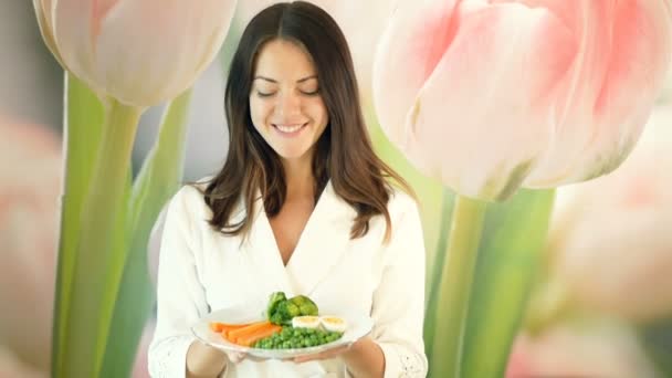 Mooi meisje in een witte robe houdt een grote plaat van gekookte groenten en vooruit duwt. Brunette knipogen. Gezonde en goede voeding. Vegetarische maaltijden. — Stockvideo