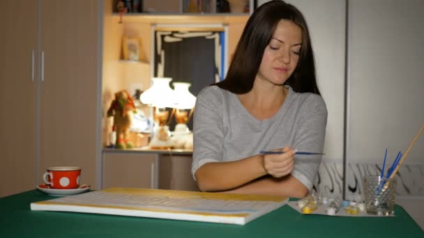 A artista iniciante pinta seu primeiro grande quadro em casa na mesa. Menina toma as mãos um pincel limpo e escolhe tinta . — Vídeo de Stock