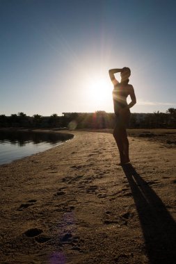 Tatlı, ince kız gün batımına karşı plaj üzerinde duruyor. Bir yüzücü bir mayo silüeti.