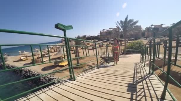 Hübsches Mädchen geht auf einer Holzbrücke im Resort und lächelt süß. Im Hintergrund ein blaues Meer und viele Sonnenschirme. — Stockvideo
