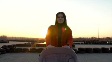 Kendine güvenen bir annenin bebek arabasıyla yürümesi. Gün batımı arkada, siluet. Bebek arabasında küçük bir bebek..