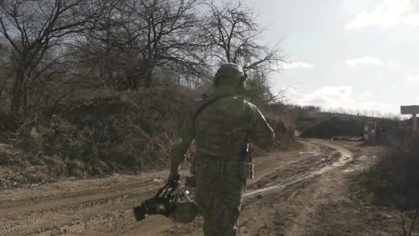 Plně vybavený neznámý voják v maskáčích kráčí špinavou cestou a nese helmu. Vojenské akce v lese. Jaro, podzim nebo zima. — Stock video
