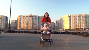 Kırmızı ceketli genç anne bebek arabasında bir bebekle koşuyor. Mutlu anne ve bebek. Dışarı çıkın. Arka planda ikamet alanı var. Aile hayatı..