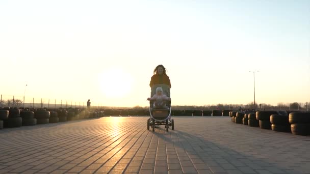 Feliz madre riendo corre con un cochecito y un bebé sonriente en ella. Tiempo libre familiar. Exterior y puesta de sol . — Vídeo de stock