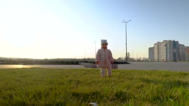 Niña con abrigo camina sobre la hierba verde. Hora del atardecer. Karting y zona residencial en el fondo. Primavera . — Vídeo de stock