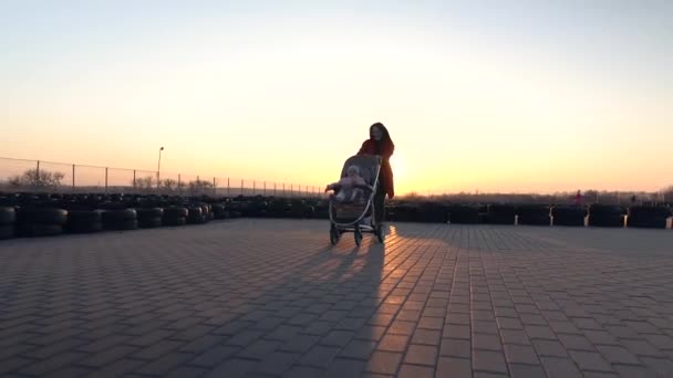 Mamá joven con abrigo rojo corre con un bebé en el cochecito. Feliz madre y niño. Puesta de sol en el fondo. Frente, costado, espalda disparada . — Vídeo de stock