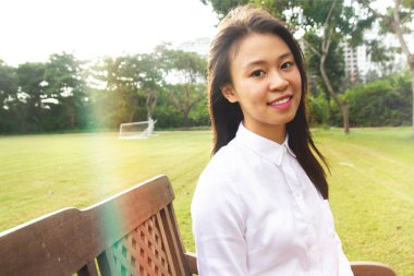 Üniversitedeki yeşil alanda güneşin altında oturmuş gülümseyen genç öğrencinin portresi.