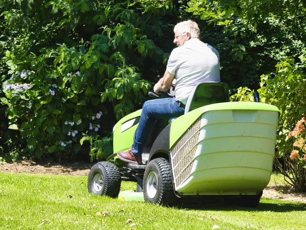 75歳の老人は 花のある庭でトラクター芝刈り機を運転しています 緑と白のパワーライド カラフルな花の間のフィールドに回転 — ストック写真