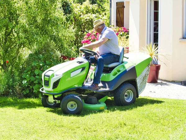 Senior man 75 old years driving a tractor lawn mower in garden with flowers. Green and white ride on mower, turning in field between colorful flowers