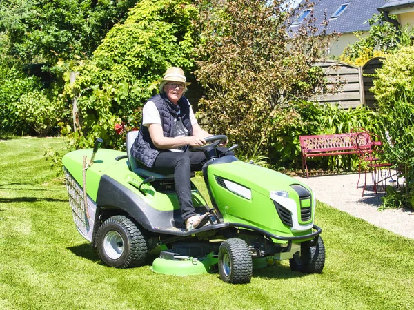 Senior man 75 old years driving a tractor lawn mower in garden with flowers. Green and white ride on mower, turning in field between colorful flowers