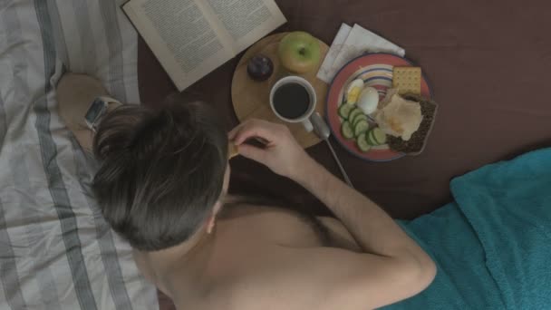 Νεαρός άνδρας τρώει νόστιμο πρωινό στο κρεβάτι. Η θέα από την κορυφή — Αρχείο Βίντεο
