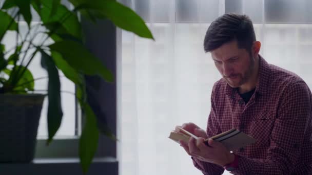 Ein Mann liest ein Buch, das am Fenster sitzt. — Stockvideo
