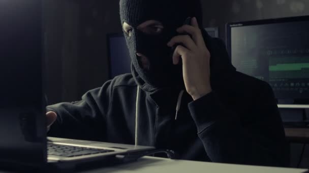 Farliga hacker i en mask talar telefon på en dator i en mörk kontorsrum — Stockvideo