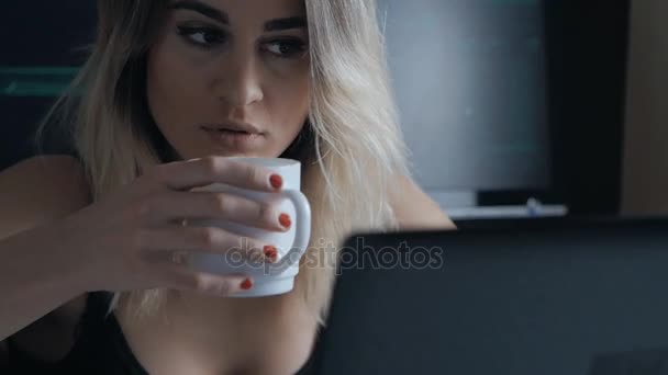 Блондинка хакер працює в ноутбуці з чашкою кави в темній кімнаті — стокове відео