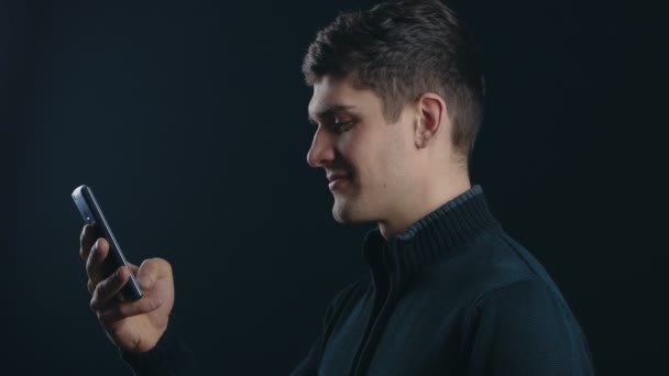 Junger, gutaussehender Mann mit Smartphone vor schwarzem Hintergrund — Stockvideo
