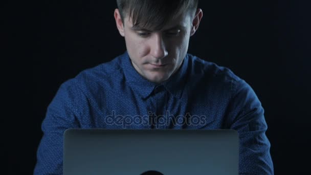 Портрет человека в синей рубашке, работающего за ноутбуком в темной комнате — стоковое видео