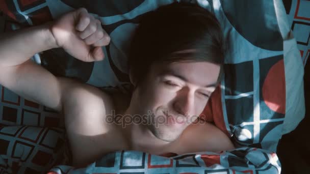 Молодой человек просыпается и растягивается в своей постели в солнечное утро — стоковое видео